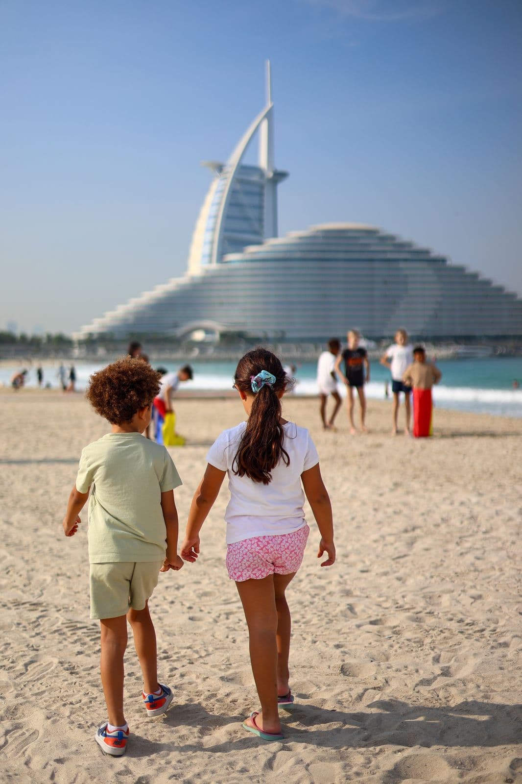 Des enfants se promènent main dans la main sur la plage, avec le Burj Al Arab en arrière-plan